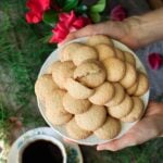 person holding a plate of gluten free ratafia biscuits with a cup of coffee and vase of roses in the background