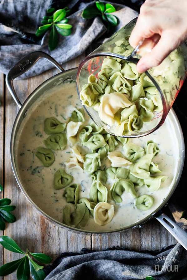 person dumping tortellini into alfredo basil sauce