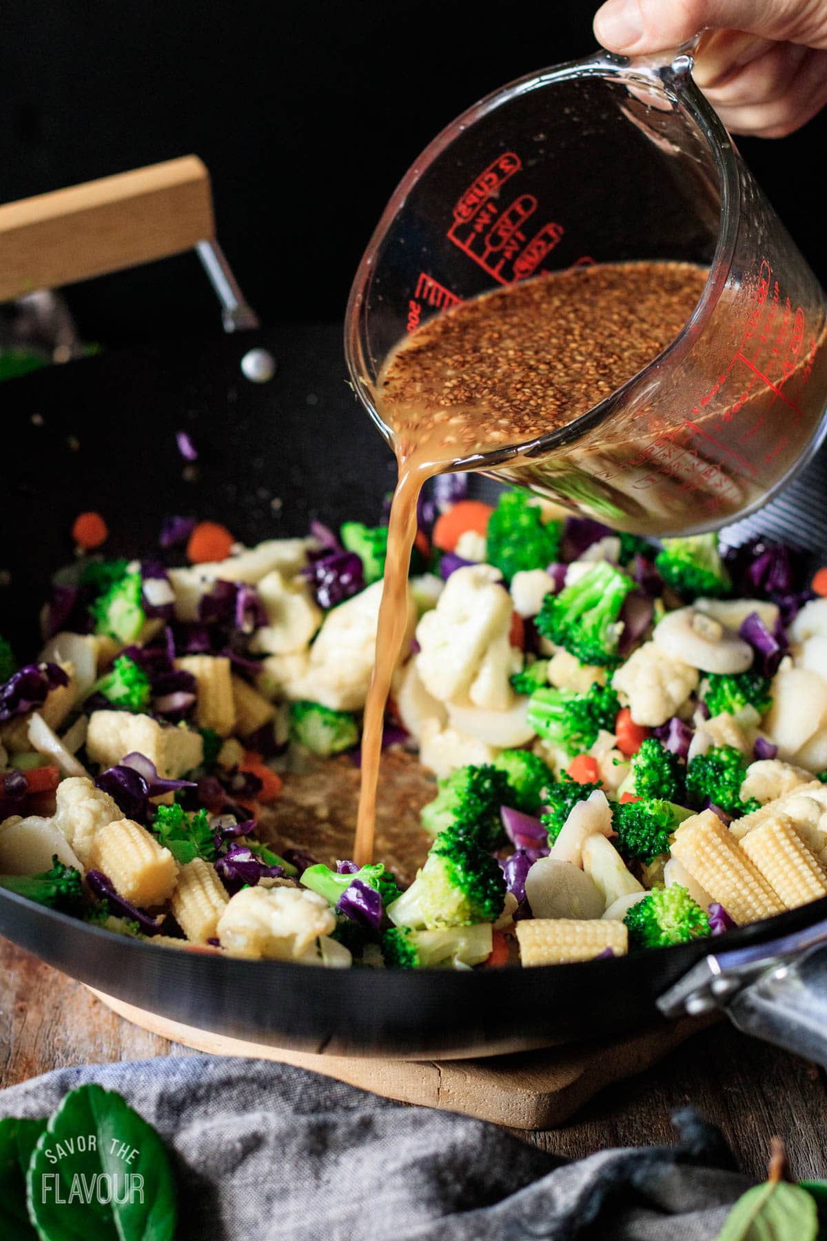 person pouring vegan stir fry sauce into a wok of cooked veggies