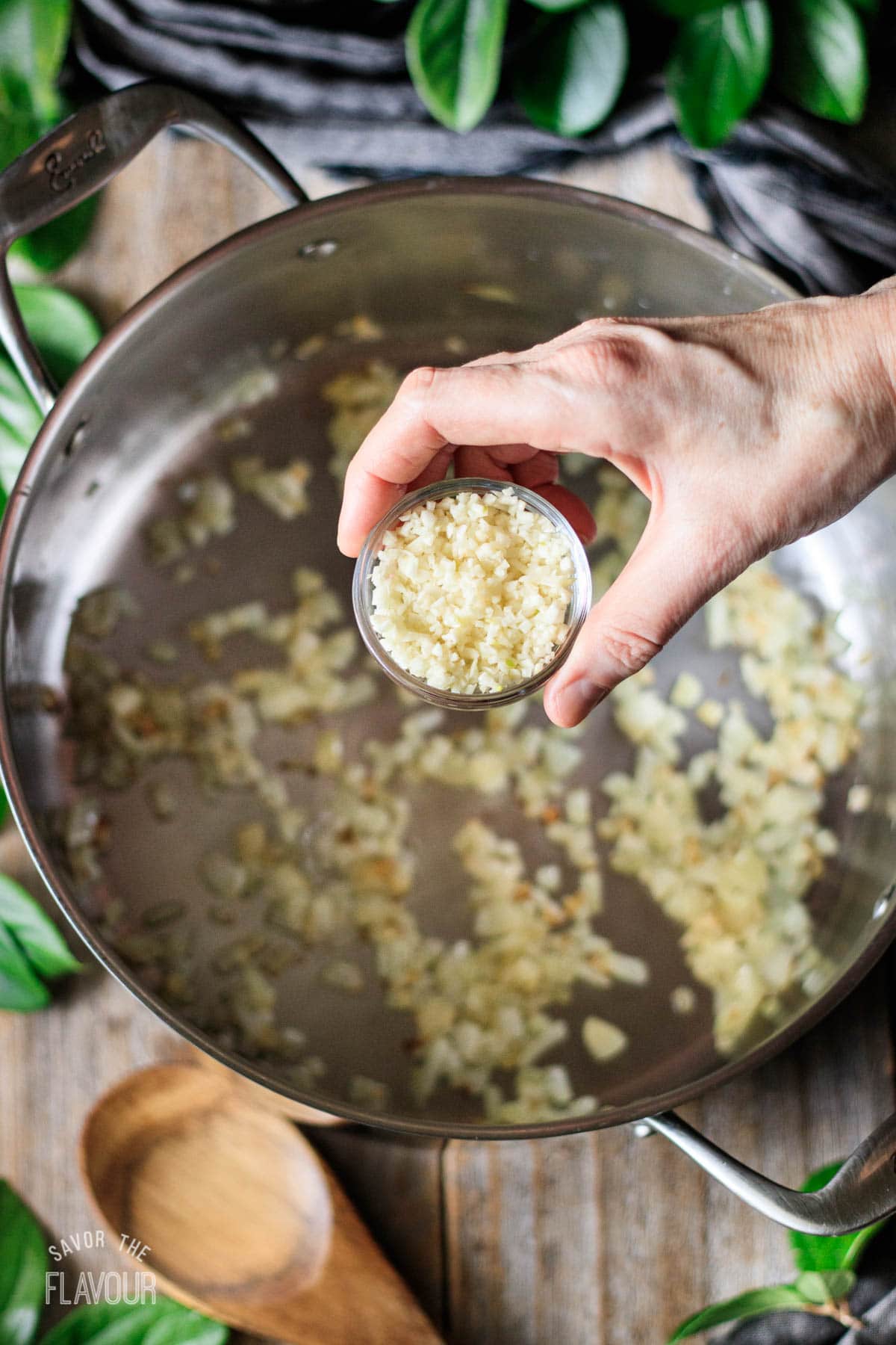 person holding a bowl of minced garlic above a pan of sauteed onion
