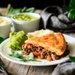 slice of minced beef pie on a white plate with peas and potatoes