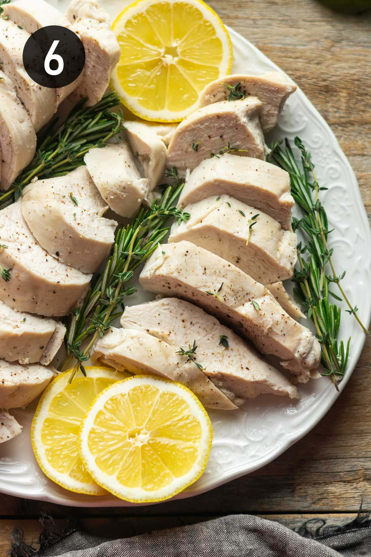 sliced poached chicken breasts on a white platter with lemon slices and fresh herbs.