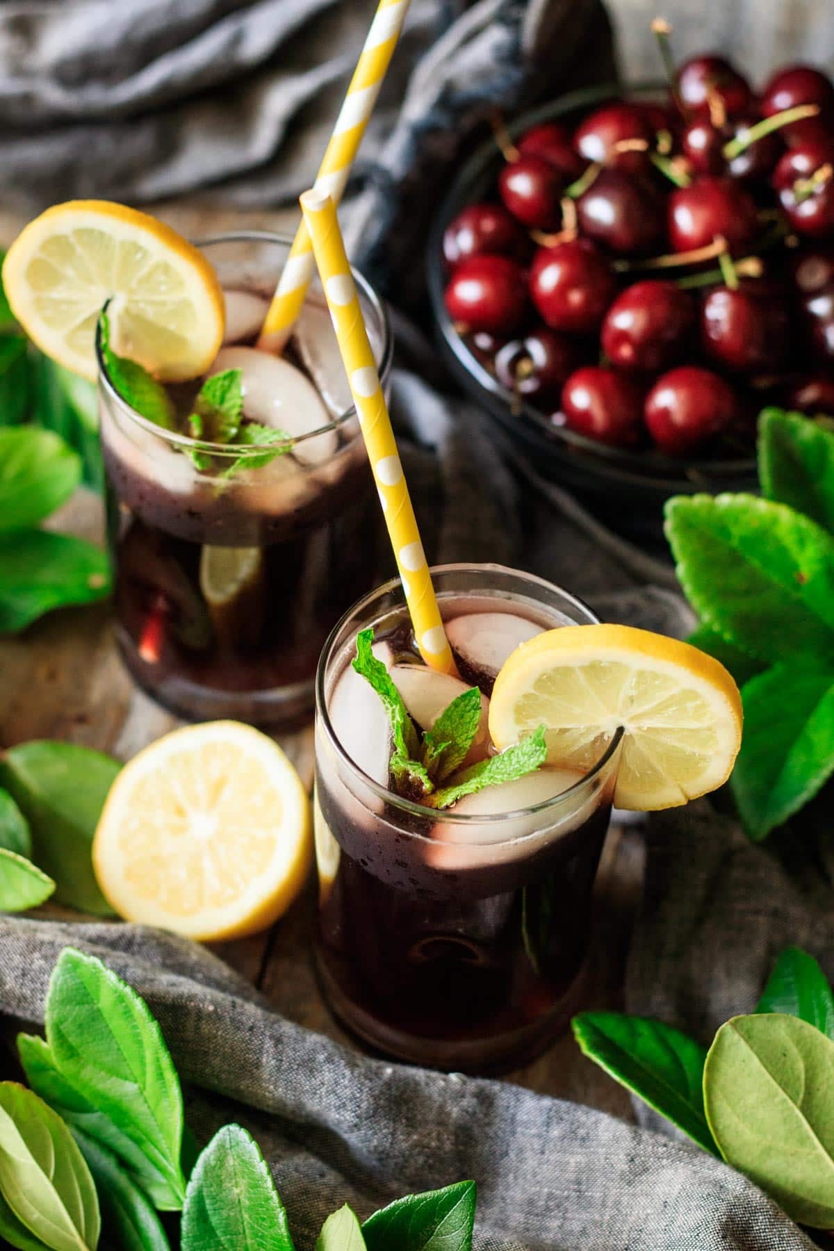 two glasses of cherry lemonade garnished with mint, lemon slices, and paper straws.
