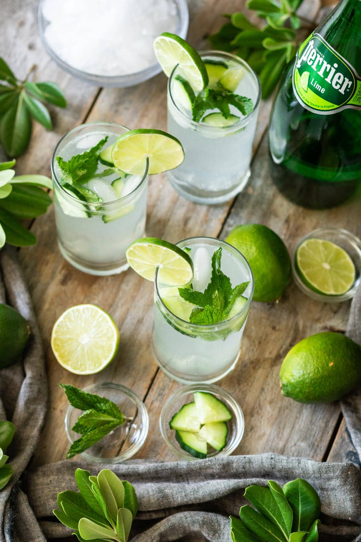 three glasses of lime and mint drink with garnishes and a bottle of Perrier.