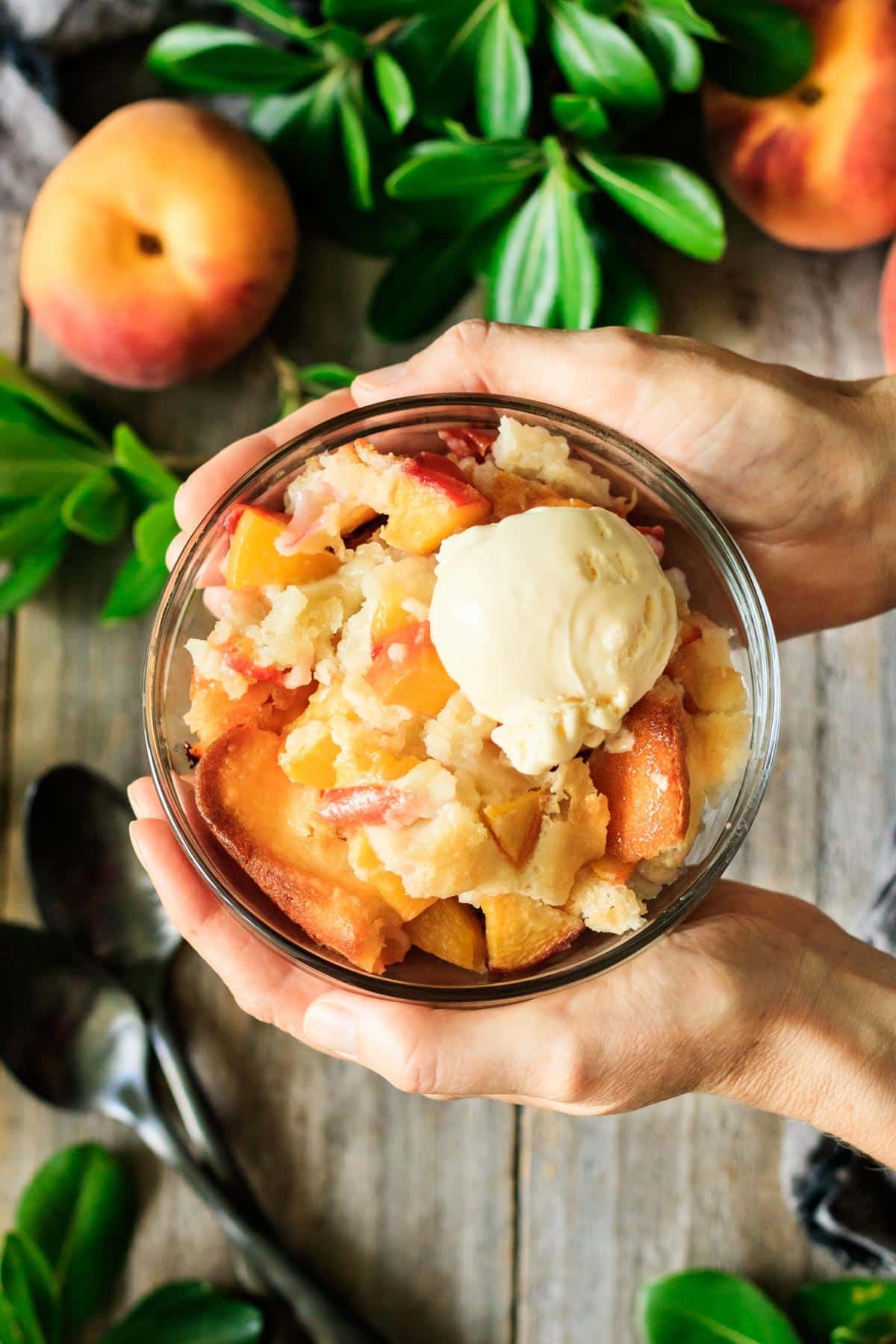 holding a glass bowl of peach cobbler with a scoop of vanilla ice cream on top.