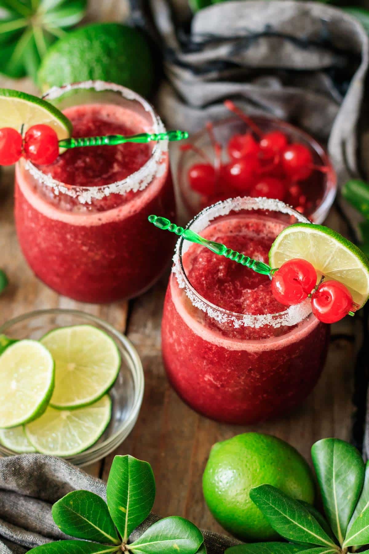 two stemless glasses of frozen cherry limeade with bowls of lime wheels and maraschino cherries.
